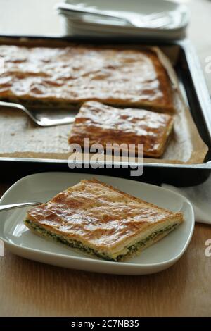 A piece of a spinach pie in puff dough served on a white plate the sheet with the rest is in the background. Stock Photo