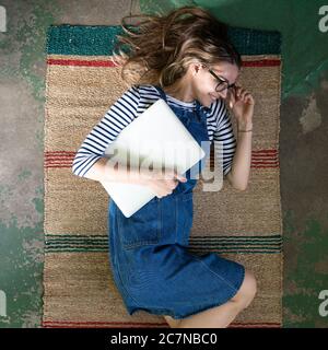 Top view of cute woman in glasses wear blue jeans overalls, lying on reed mat on the concrete floor, holding laptop and resting after work. Stock Photo