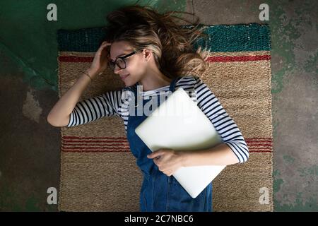 Top view of cute woman in glasses wear blue jeans overalls, lying on reed mat on the concrete floor, holding laptop and resting after work. Stock Photo