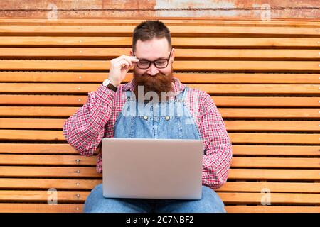 Close up portrait of pleasant brutal bearded hipster man in blue overalls, checked shirt working on laptop sitting on bench outdoors. Distance job Stock Photo