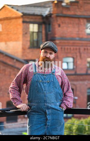 Portrait of brutal bearded man wearing blue overalls, checked shirt and cap in vintage style of the mid 20th century, looking at camera, outdoors. Stock Photo