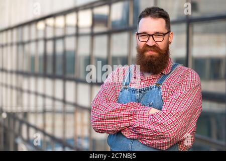 Portrait of brutal bearded stylish craftsman wearing blue overalls, checked shirt in vintage style of the mid 20th century, looking at camera, outdoor Stock Photo