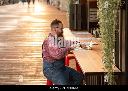 Portrait of brutal bearded hipster man freelancer wearing blue overalls and checked shirt working on laptop sitting in cafe/restaurant outdoors. Side Stock Photo