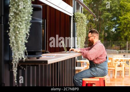 Portrait of brutal bearded hipster man freelancer wearing blue overalls and checked shirt working on laptop sitting in cafe/restaurant outdoors. Side Stock Photo