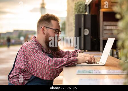 Portrait of brutal bearded hipster man freelancer wearing blue overalls and checked shirt working on laptop sitting in cafe/restaurant outdoors. Side Stock Photo
