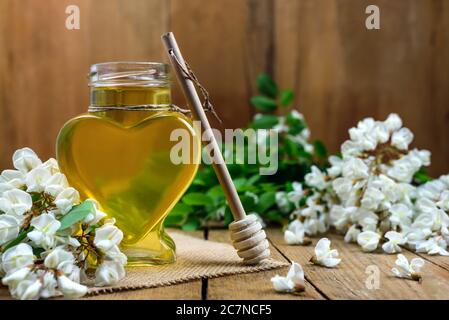 Sweet honey in a heart-shaped glass jar with a wooden dipper and acacia flowers on the old table with copy space for text. Selective focus. Stock Photo