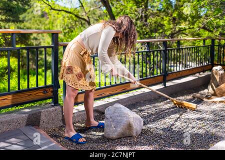 Zen rock garden outside in Japan with woman holding rake raking stone making pattern on gravel with fence and green foliage background Stock Photo