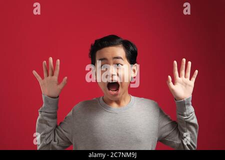 Portrait of funny young Asian boy looking at camera with open mouth and big eyes, shocked surprised expression against red background Stock Photo