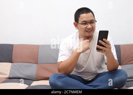 Happy smiling Asian man using smartphone device while sitting on sofa at home. Male reading messages on mobile phone while relaxing at home Stock Photo