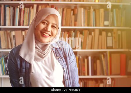 Portrait of cheerful Asian muslim female librarian wearing hijab, looking at camera and smiling, woman standing against books in library Stock Photo