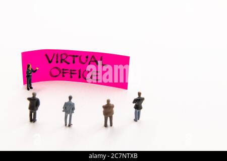 Strategy meeting with miniature figurines posed as business people standing around post-it note with Virtual Office handwritten message on white backg Stock Photo