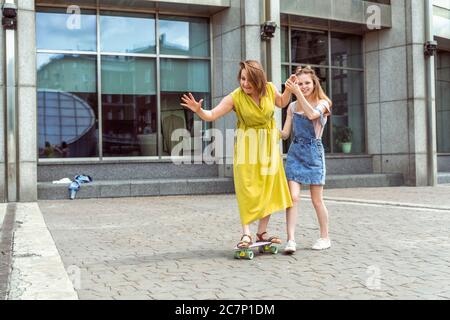 Young mother practising on skateboard in the city street. Daughter helping mum on skateboard. Mum learning to ride skateboard as Daughter teaches her Stock Photo