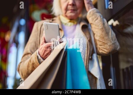 Sad elderly female standing on the street while waiting call on her smartphone Stock Photo