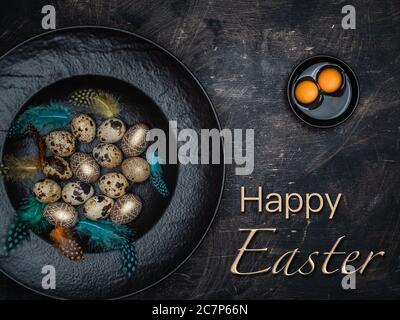 Easter concept with text: Happy Easter. Natural quail eggs, golden colored eggs and colorful feathers in black bowl. Stock Photo
