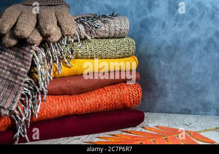 Stack of folded warm knitted women's sweaters in warm colors, plaid scarf, knitted gloves and autumn leaves on gray background. Copy space. Closeup Stock Photo