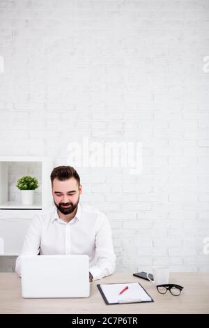 portrait of cheerful bearded businessman using laptop in office - copy space over white brick wall background Stock Photo