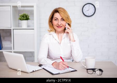 cheerful mature business woman sitting in modern office, talking by phone and writing something on clipboard Stock Photo