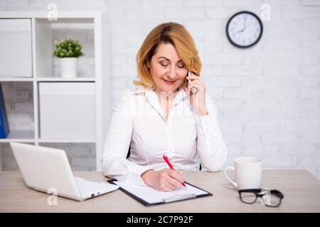 portrait of mature business woman sitting in modern office, talking by phone and writing something on clipboard Stock Photo