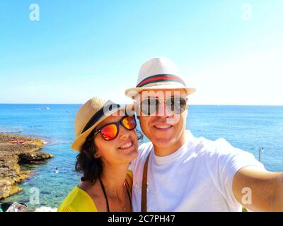 Shallow focus shot of a smiling couple wearing hat and sunglasses taking selfies at the beach Stock Photo