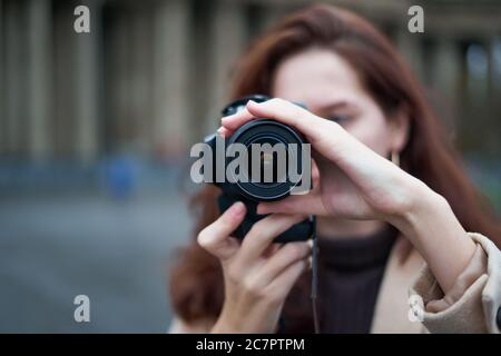 Selective focus on lens. Beautiful stylish fashionable girl holds camera in her hands and takes pictures. Woman photographer with long dark hair in Stock Photo