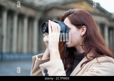 Beautiful stylish fashionable girl holds camera in her hands and takes pictures. Woman photographer with long dark hair in city, urban shoot Stock Photo
