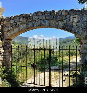 Rusty metal gate in the stone wall at the old medieval farmhouse in Provence, dirt road along countryside on background. France Stock Photo