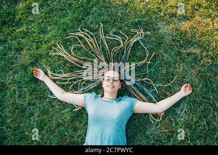 Happy young woman is lying on grass with box braids arranged like sun enjoying summer days. Stock Photo