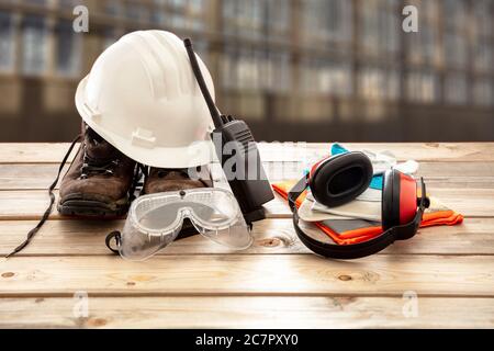 Work safety protection equipment. Industrial protective gear on wooden table, blur construction site background. Hardhat, boots, earmuffs, walkie talk Stock Photo