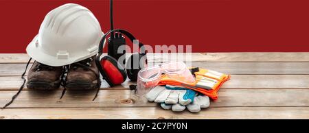 Work safety protection equipment. Industrial protective gear on wooden table, red color background. Hardhat, boots, earmuffs, walkie talkie, glasses a Stock Photo