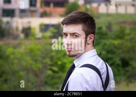 Portrait of stylish brunet young man, wearing white shirt, black tie, suspenders and glasses, walking on the meadow near river. Stock Photo