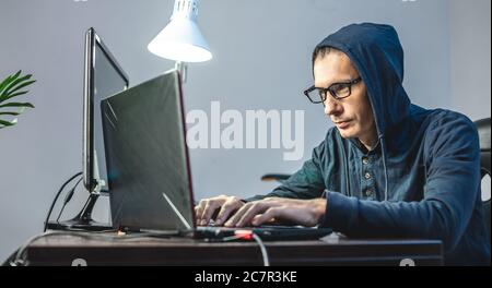Man hacker in the hood is programming virus code at the keyboard for the laptop. The concept of Internet fraud and personal data hacking Stock Photo