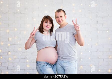 cute pregnant couple showing victory sign over white brick wall Stock Photo