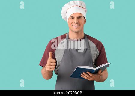 Young man with recipe book showing thumb-up gesture on color background Stock Photo