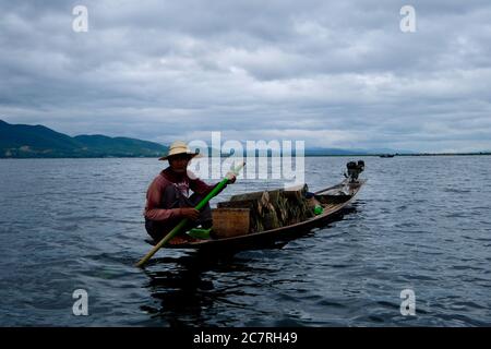 close up one middle-aged fisherman sitting on boat in Inle lake, Myanmar. Rowing the boat looking at camera. White cloudy sky with mountains. Stock Photo