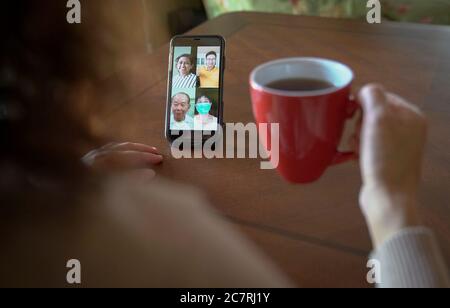 Woman with a cup of coffee having a group videocall and chat over her mobile phone at home Stock Photo