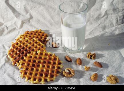 A glass of milk, Viennese waffles and walnuts on a white tablecloth, taken with harsh light. Stock Photo