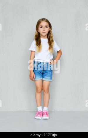 Little girl in white t-shirt, jeans shorts and pink sneakers standing with hand in pocket near the wall Stock Photo