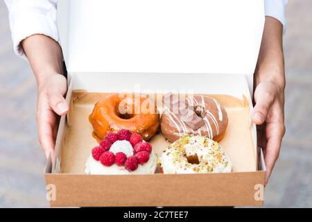 close up hands holding four pieces of totally different colorful and delicious looking donuts in ecological carton box. appetising fast food dessert Stock Photo