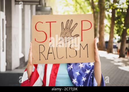 Woman with flag of USA holding placard with text STOP RACISM outdoors Stock Photo