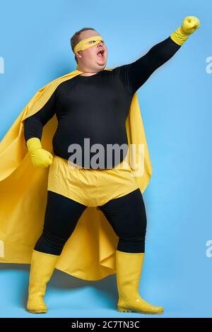 handsome brave strong man has a mad desire, a dream to shield the world from dirt and evil. fat superhero in yellow costume posing isolated over blue Stock Photo