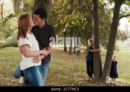 Young couple kissing while their son and daughter watching them hiding behind a tree in the park Stock Photo