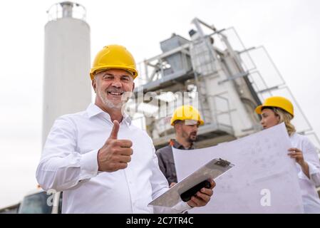 Senior engineer smiling holding a thumb up with colleagues in the background at a construction site Stock Photo
