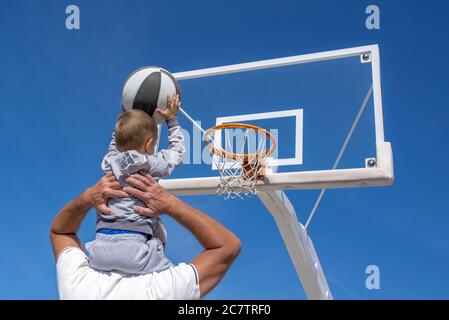 Back view of a grandson sitting on his grandfather's shoulder throwing a basketball into a hoop Stock Photo