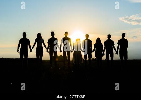 Cohesion concept. Black silhouettes of friends holding hands stands at sunset. Stock Photo