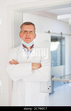 Mature smiling doctor with face mask standing confidently in front of the surgery room - focus on the face Stock Photo