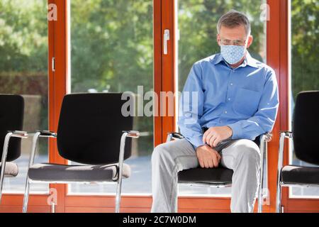 Sad or tired mature man with face mask sitting in a waiting room of  a hospital or an office on a sunny day Stock Photo