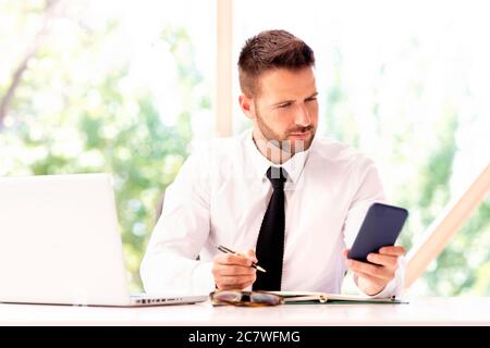 Shot of financial businessman making notes and text messaging while sitting at office desk and working. Stock Photo