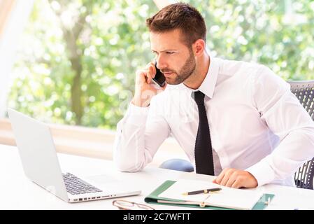 Shot of confused businessman wearing shirt and tie while sitting at office desk and working. Professional man talking with somebody on his mobile phon Stock Photo