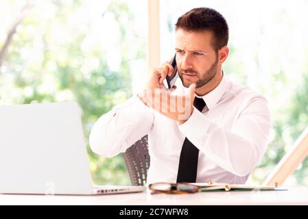 Shot of confused businessman wearing shirt and tie while sitting at office desk and working. Professional man talking with somebody on his mobile phon Stock Photo