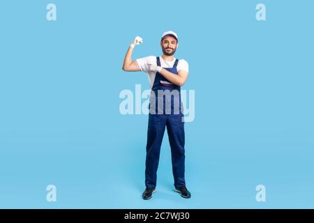 Full length happy expert handyman in blue overalls, cap and protective gloves pointing biceps, demonstrating strength. Profession of service industry, Stock Photo